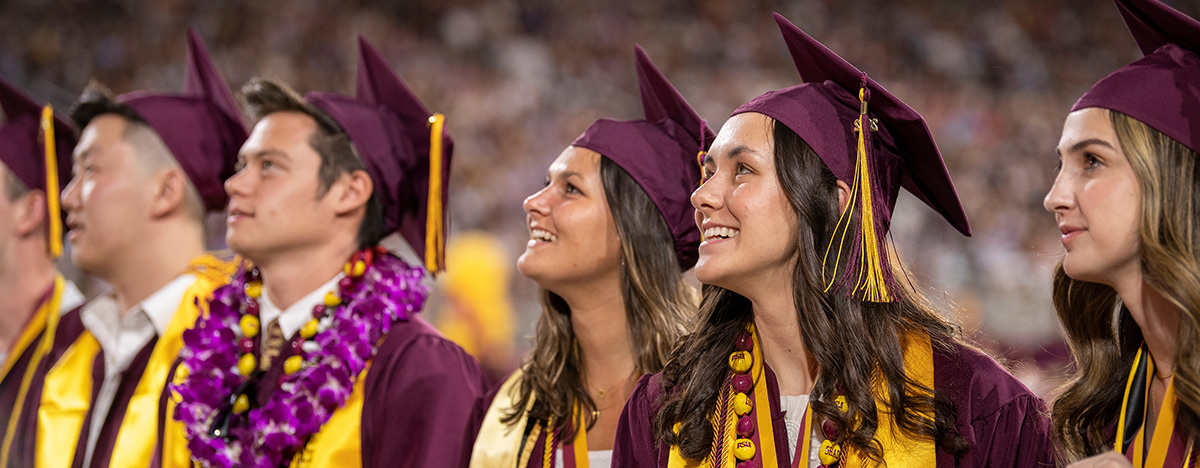 Close-up landscape photograph perspective of six ASU W.P. Carey School of Business graduating students smiling in an elated manner dressed in their maroon gowns and caps with maroon/gold colored tassels plus gold colored honor ribbons on their shoulders alongside custom necklaces/hawaiian style leis