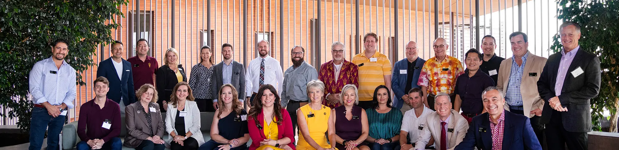 Outdoor landscape photograph of past and current W.P. Carey Sun Devil 100 winner individuals all smiling and posing for a picture as some are sitting on a couch and others are standing nearby a building area and outside planters