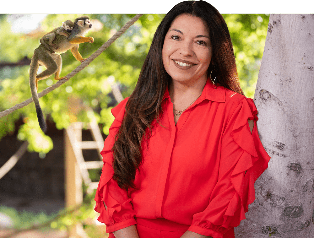 Bonnie Mendoza, wearing a bright red ruffle sleeved blouse, smiles while leaning against a tree. Behind her a small primate travels along a hanging rope while straddling a baby primate on its back.