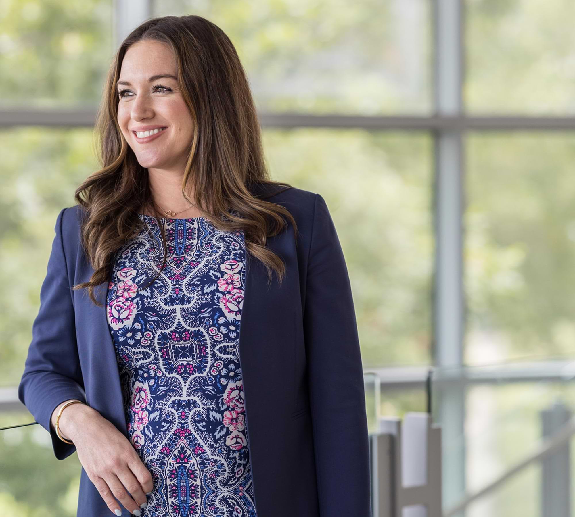 Heather Bronson, wearing a blue and pink paisley-like patterned dress under a blue blazer. She looks beyond the camera and smiles while resting her arm on a glass balcony.