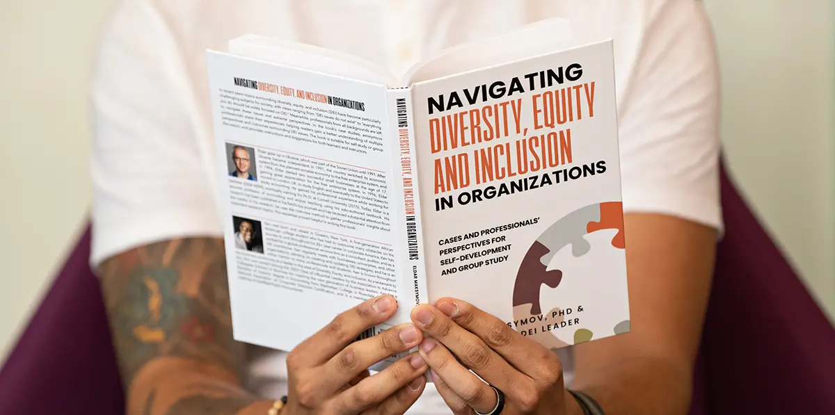 Close-up photograph perspective of a person seated down in a small dark burgundy chair with a white open book in his fingers with the front cover reading "Navigating Diversity, Equity, and Inclusion in Organizations: Cases and Professionals' Perspectives for Self-Development and Group Study"