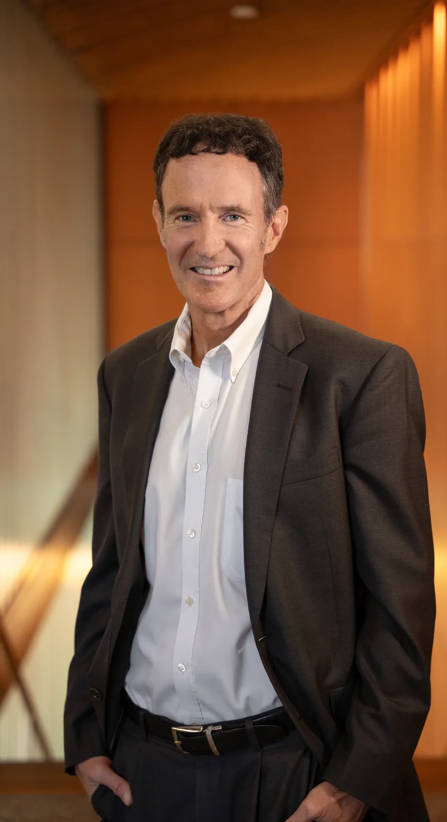 Craig Carter wearing a white button up shirt and brown blazer with his hands in his pants pockets and smiling at the camera