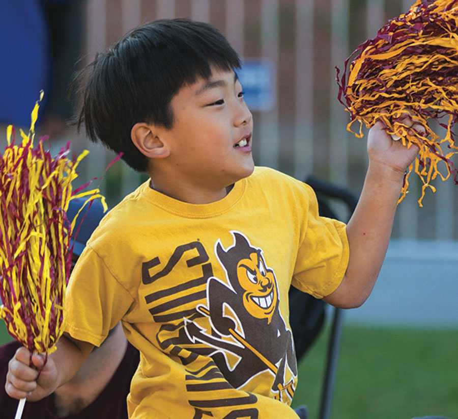 a young boy wearing a Sun Devils t-shirt waves yellow and gold shaker poms in either hand