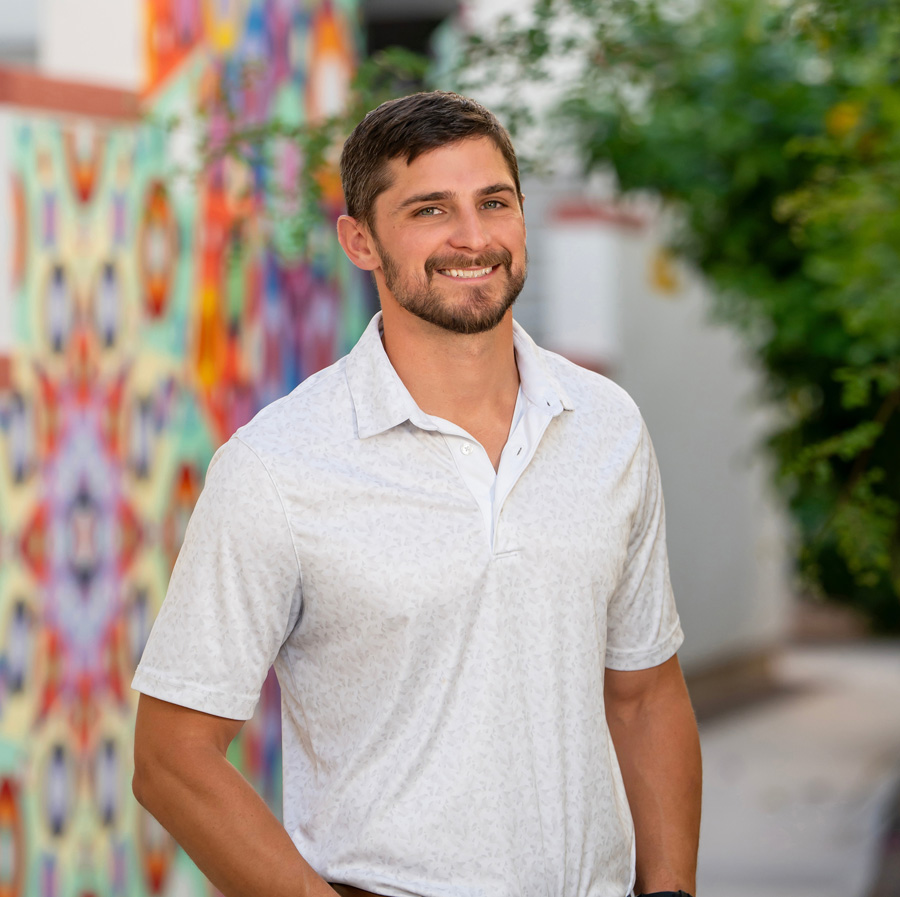 A smiling, bearded man, Rob Maloney, stands outdoors in front of a colorful, patterned mural. He wears a light-colored, subtly patterned polo shirt and a brown leather belt. The background mural features a kaleidoscope design in bright colors like pink, blue, and green.