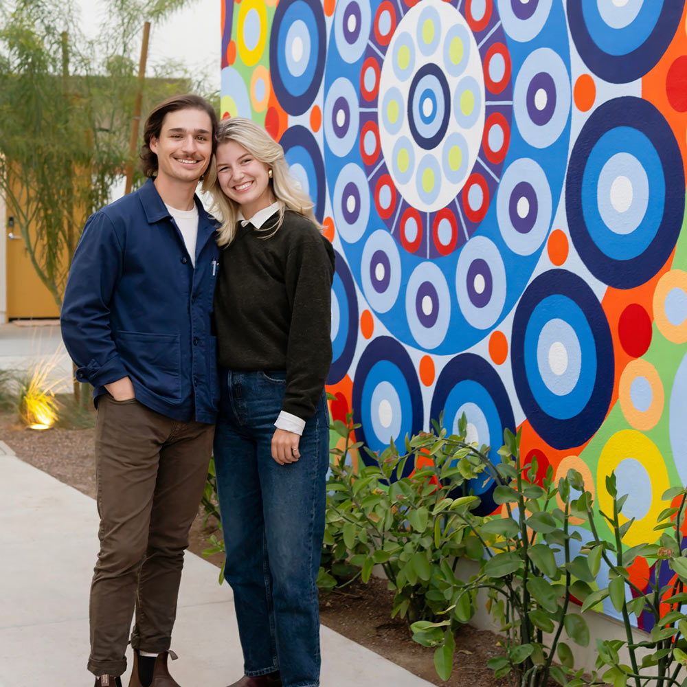 A young, smiling couple, Brad Biehl and a woman, stand outdoors in front of a brightly colored geometric mural of concentric circles in shades of blue, red, and yellow. Brad, on the left, wears a blue jacket and dark pants, while the woman wears a dark sweater over a white collar and blue jeans. Green foliage is visible in the bottom foreground.