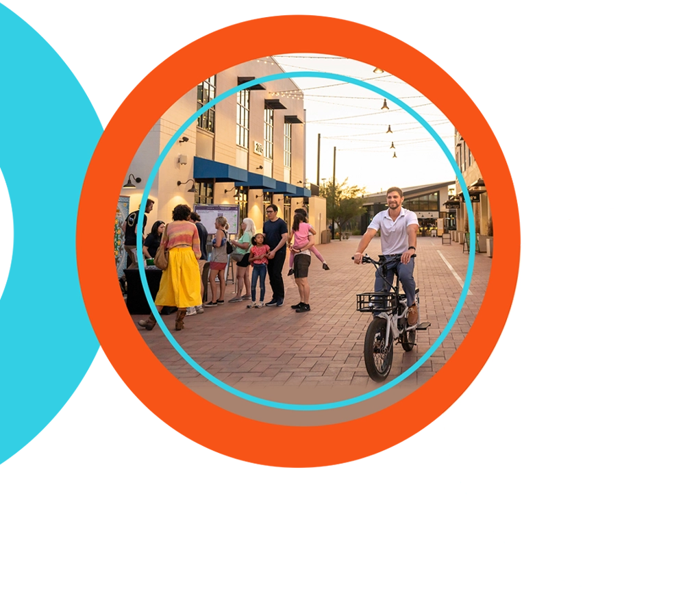 Rob Maloney is shown smiling while riding a white e-bike with a front basket down a brick-paved, pedestrian street. In the background to the left, a small group of people gathers near a table in front of a modern commercial building during the golden hour. The street is lined with businesses and illuminated by overhead string lights.