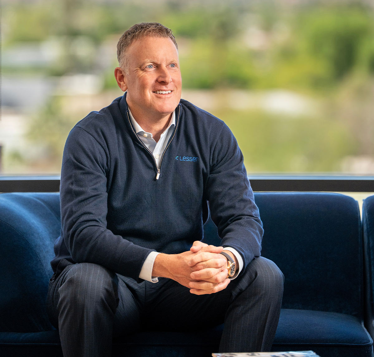 A portrait of Jay McKee, a middle-aged man with short brown hair, sitting on a deep blue velvet sofa. He is wearing a navy blue quarter-zip pullover with the logo