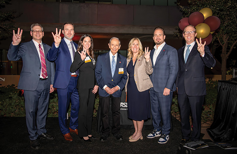 Seven people standing on a stage posing and holding up three fingers.