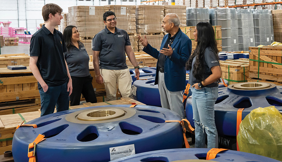 A group of people conversing near large industrial parts inside a warehouse setting.