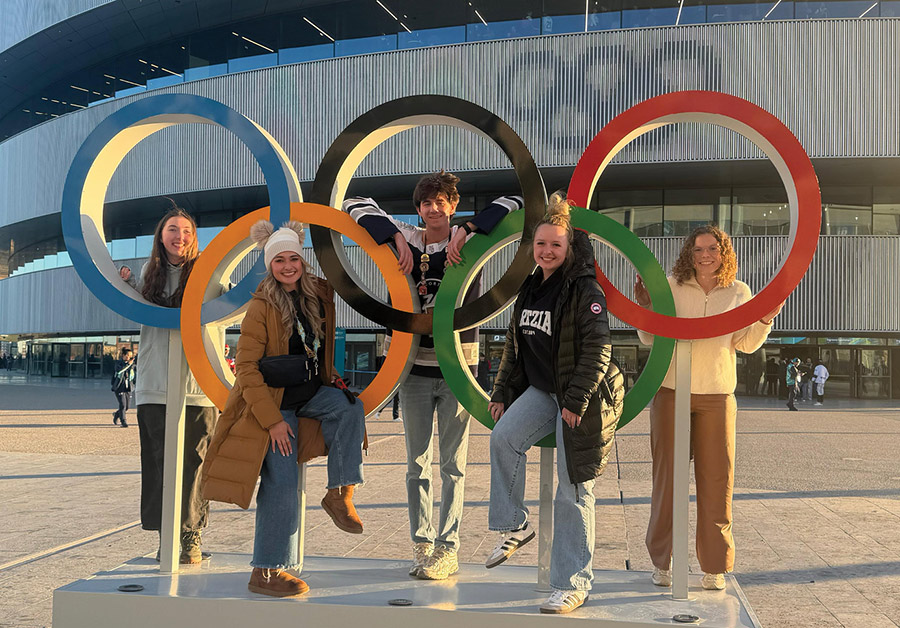 Five smiling young adults posing behind a large outdoor sculpture of the Olympic rings.